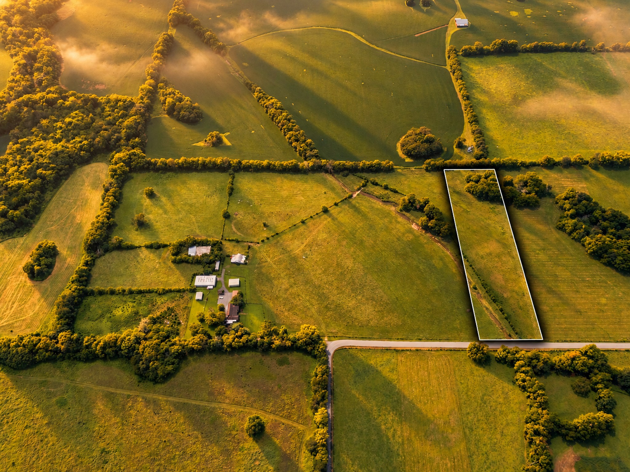 Aerial view of a long rural parcel outlined in white beside a road and tree-lined fields.