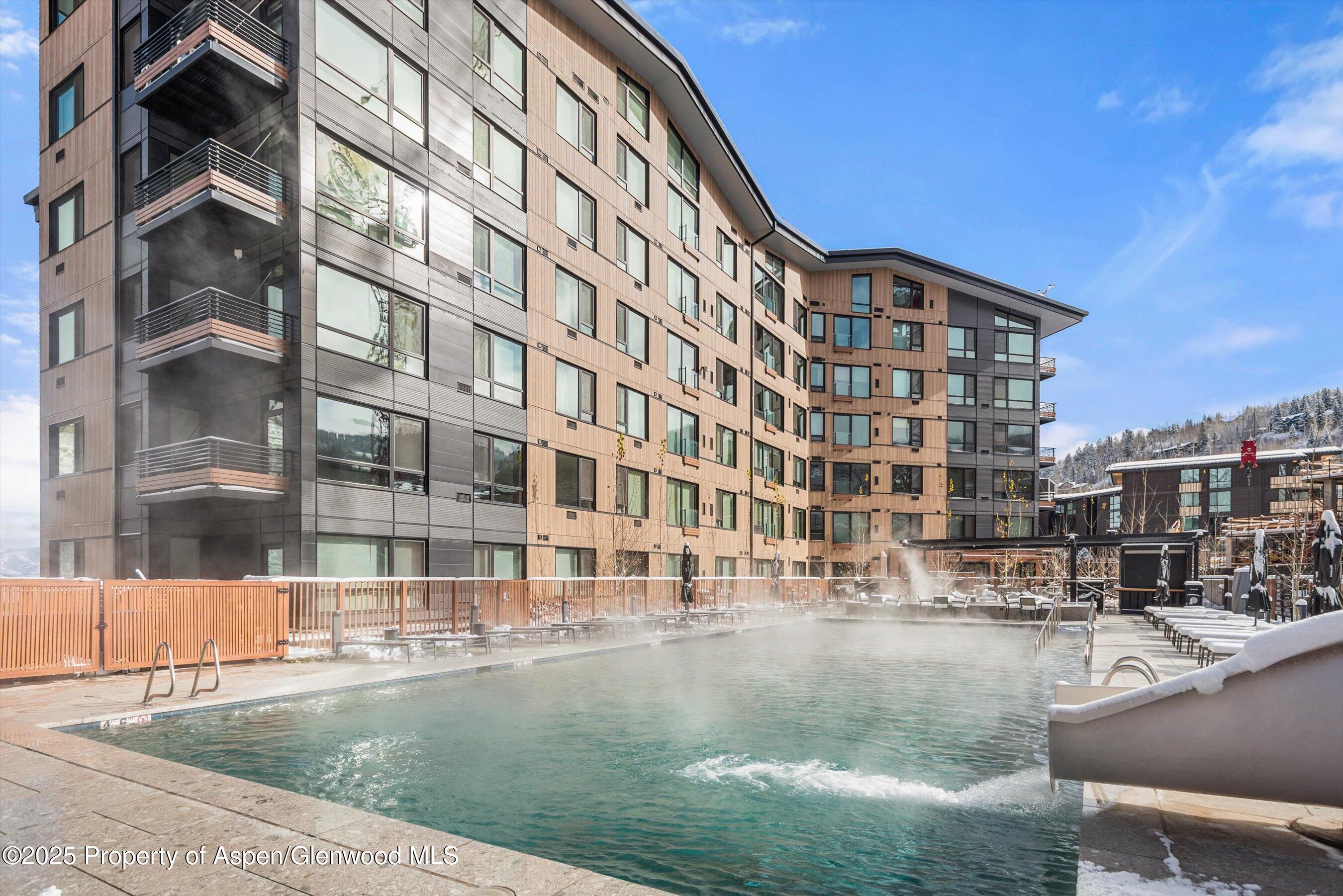 Outdoor pool steaming beside a modern condominium building, with snow on the deck and surrounding roofs.