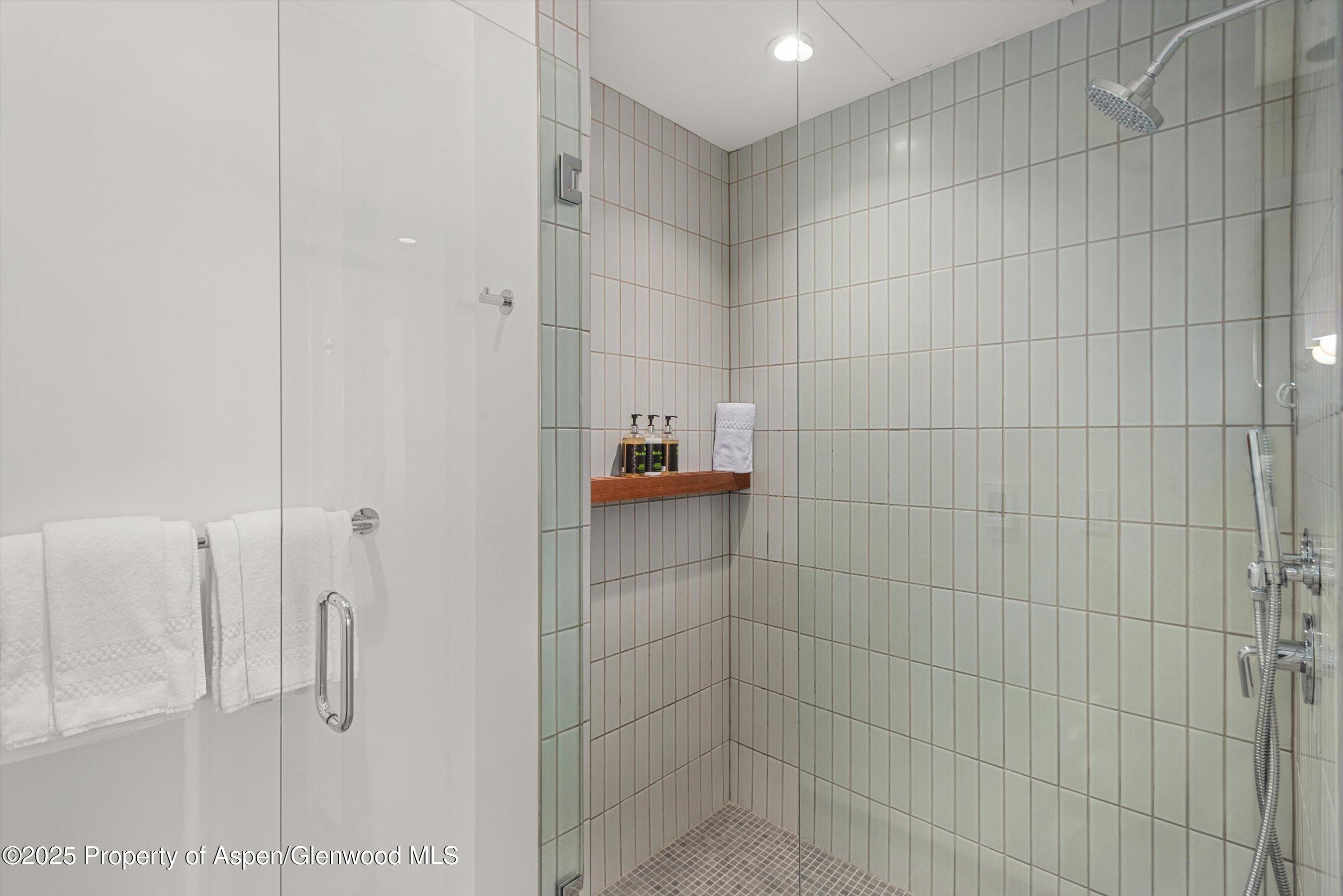 Walk-in shower with pale square tiles, chrome fixtures, and a wooden shelf holding toiletries and a towel.