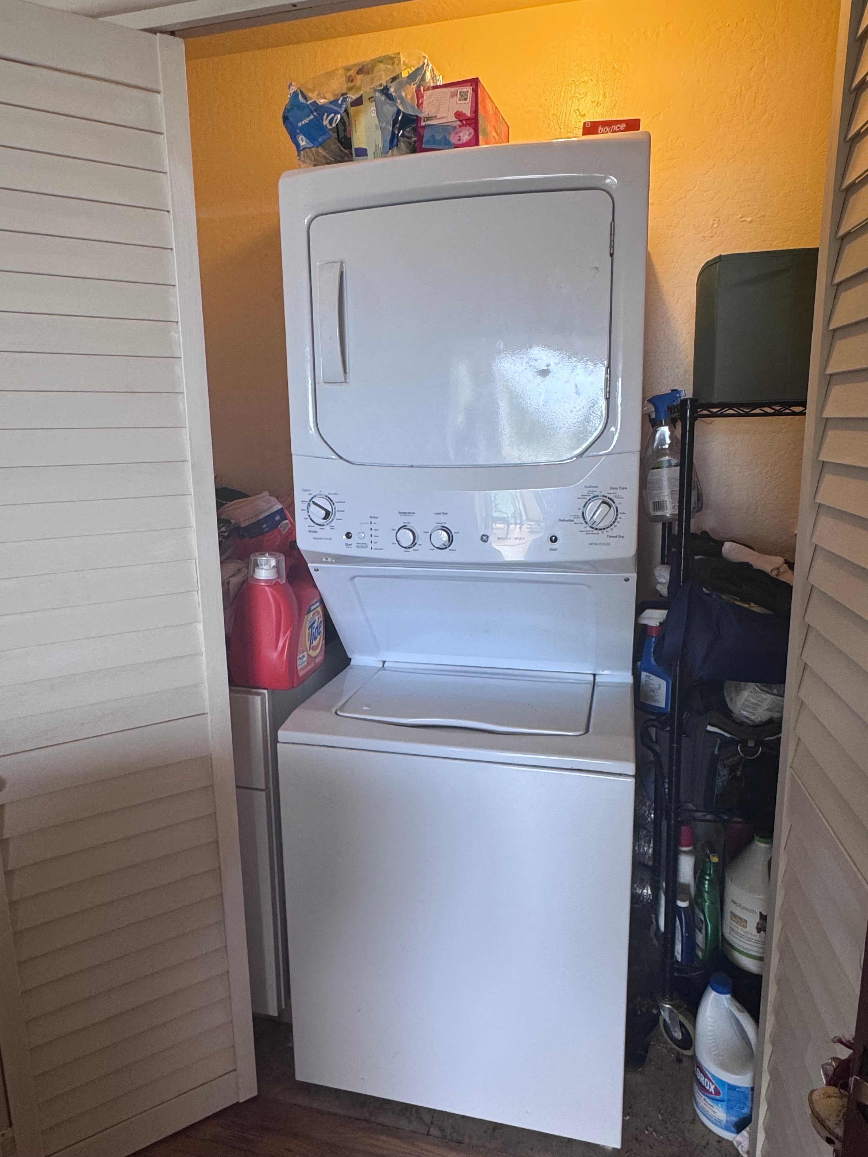 Stacked washer and dryer in a compact laundry closet with cleaning supplies and louvered doors.