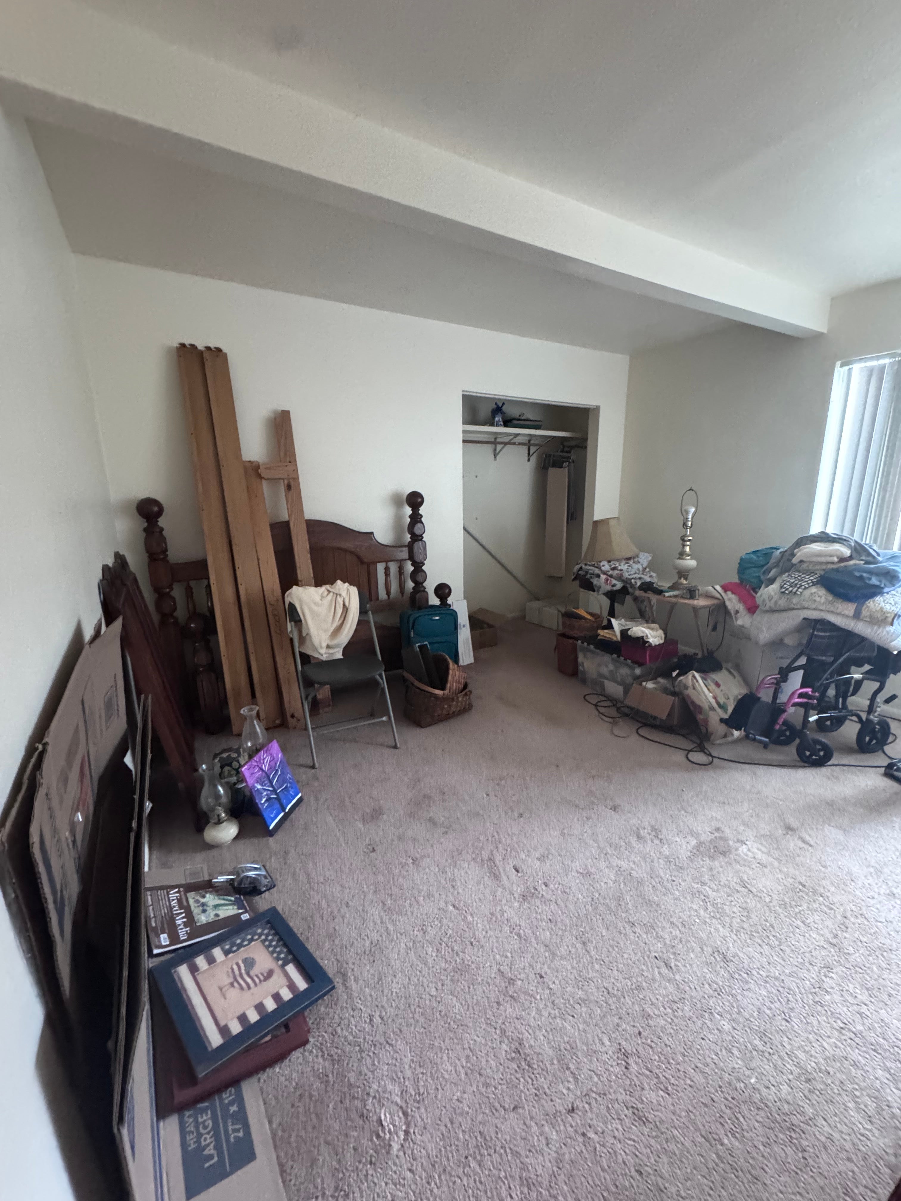 Cluttered bedroom with an open closet, disassembled bed frame, and belongings stacked beside a window.