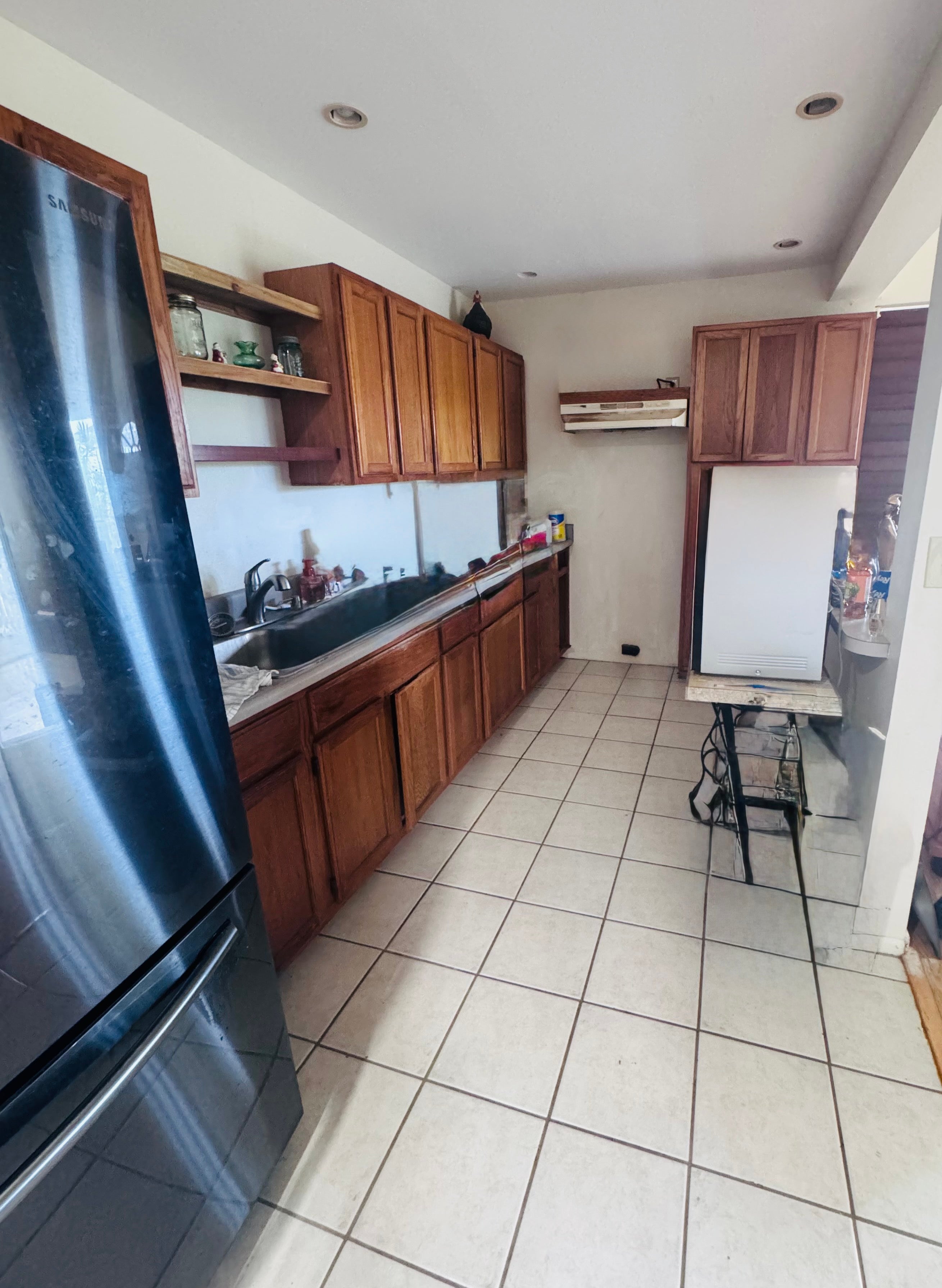 Narrow kitchen with reddish-brown cabinets, dark countertop, large refrigerator, and square tile floor.