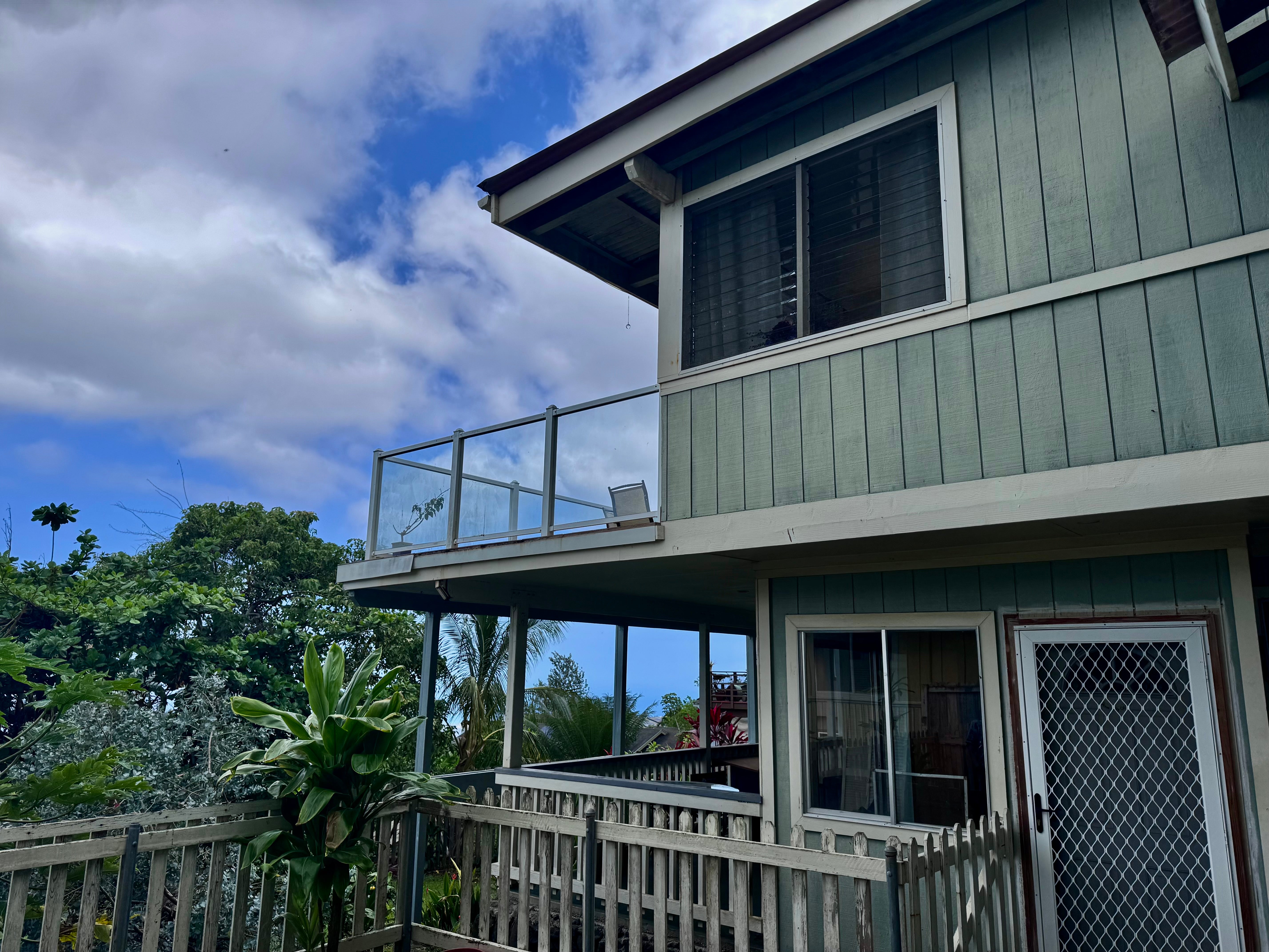 Green two-story home with a glass-railed balcony and lush tropical landscaping.