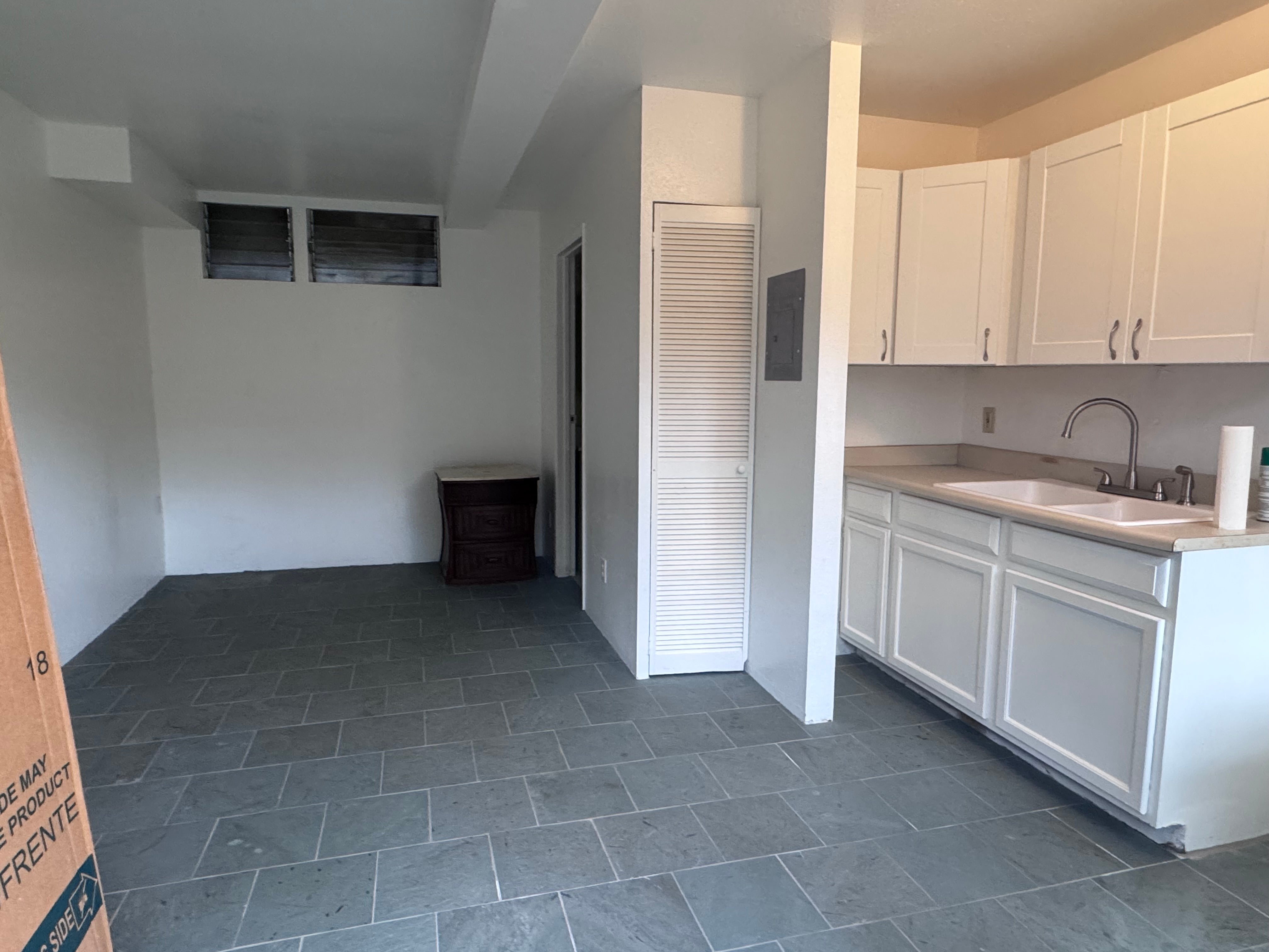 Kitchen with white cabinets and double sink beside an empty room with dark gray tile flooring.