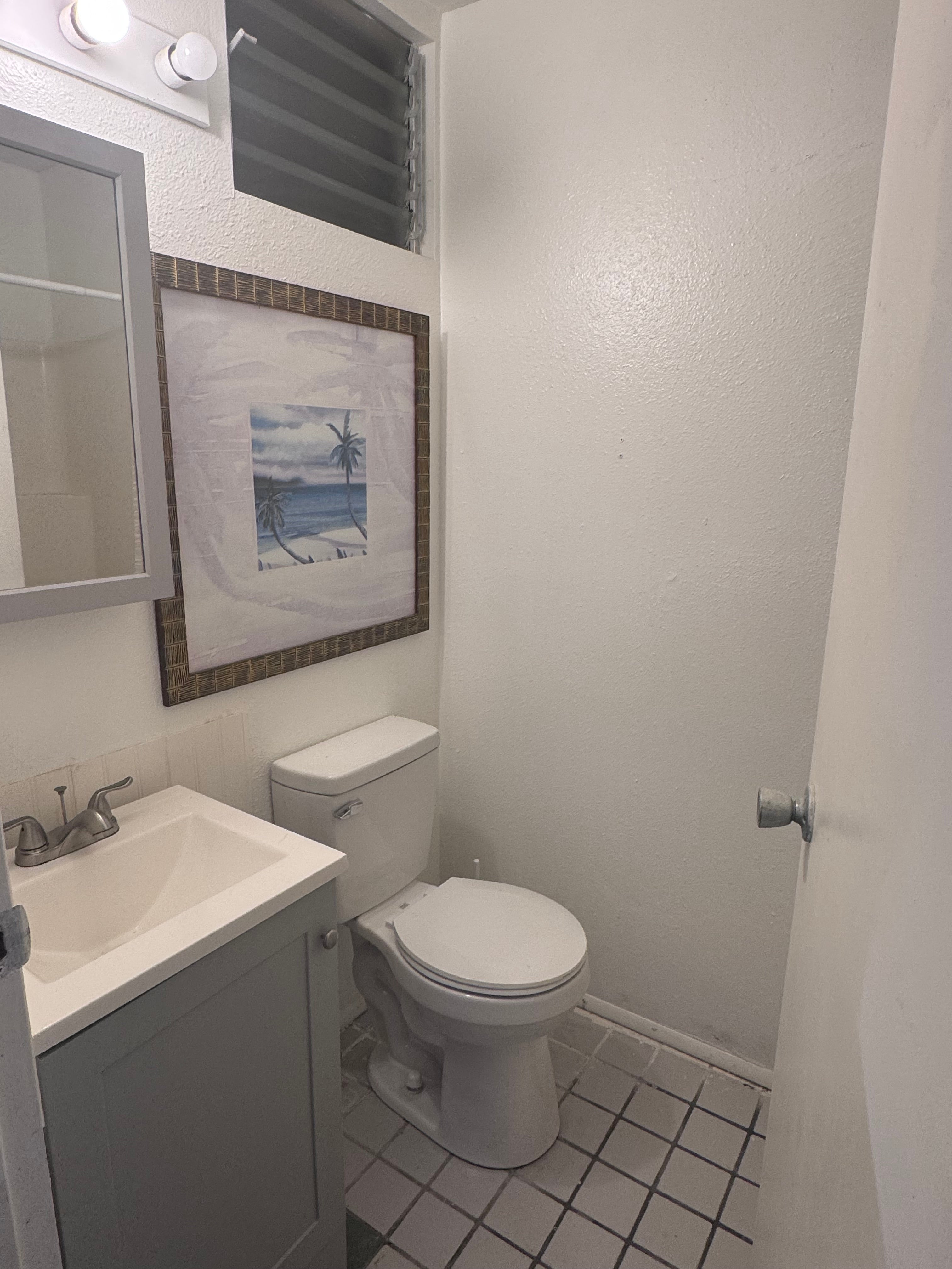 Compact bathroom with a toilet, gray vanity, tiled floor, and framed tropical beach artwork.