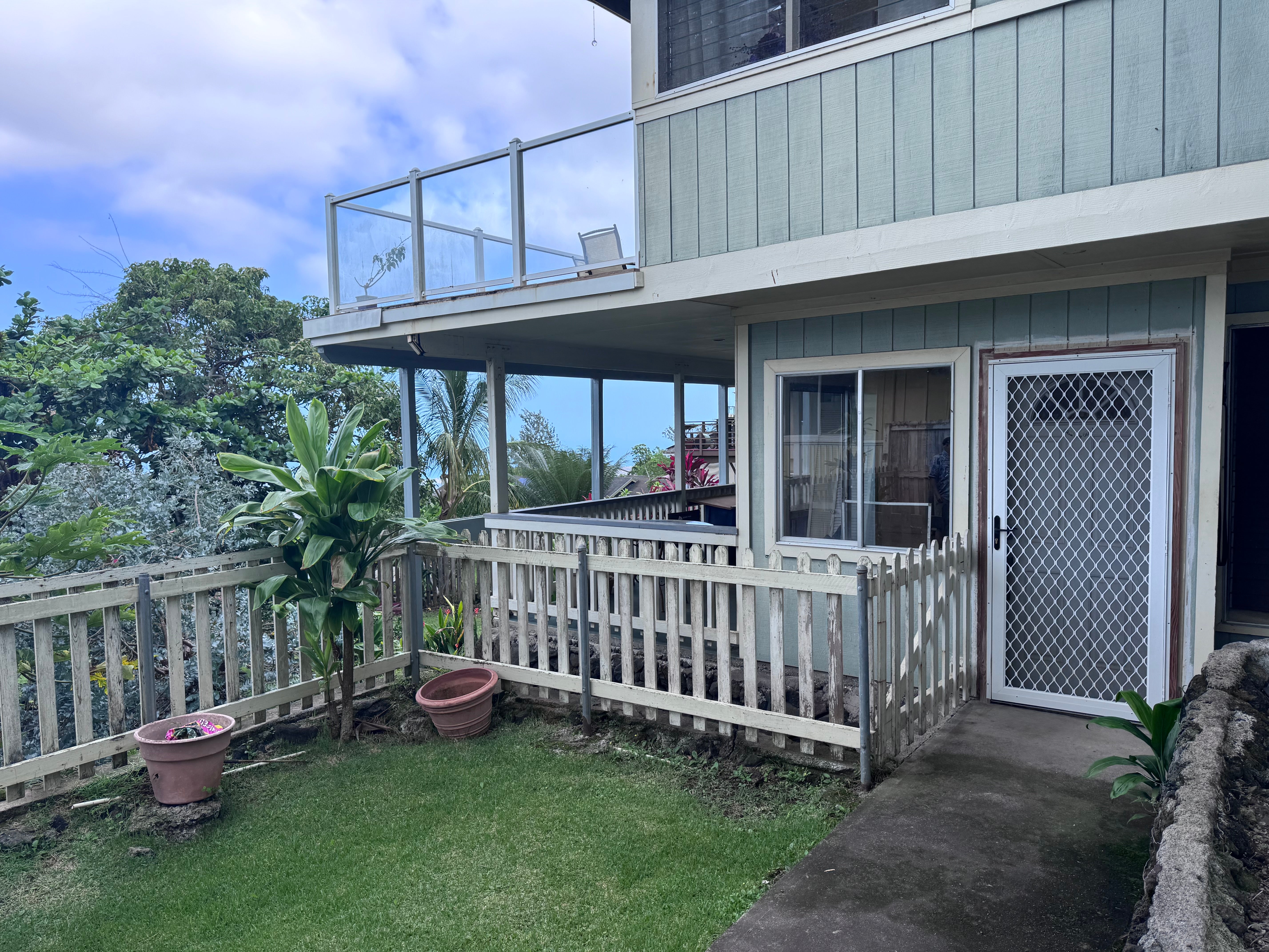Two-story blue-gray home with an upper balcony, screened entry, and fenced tropical garden.
