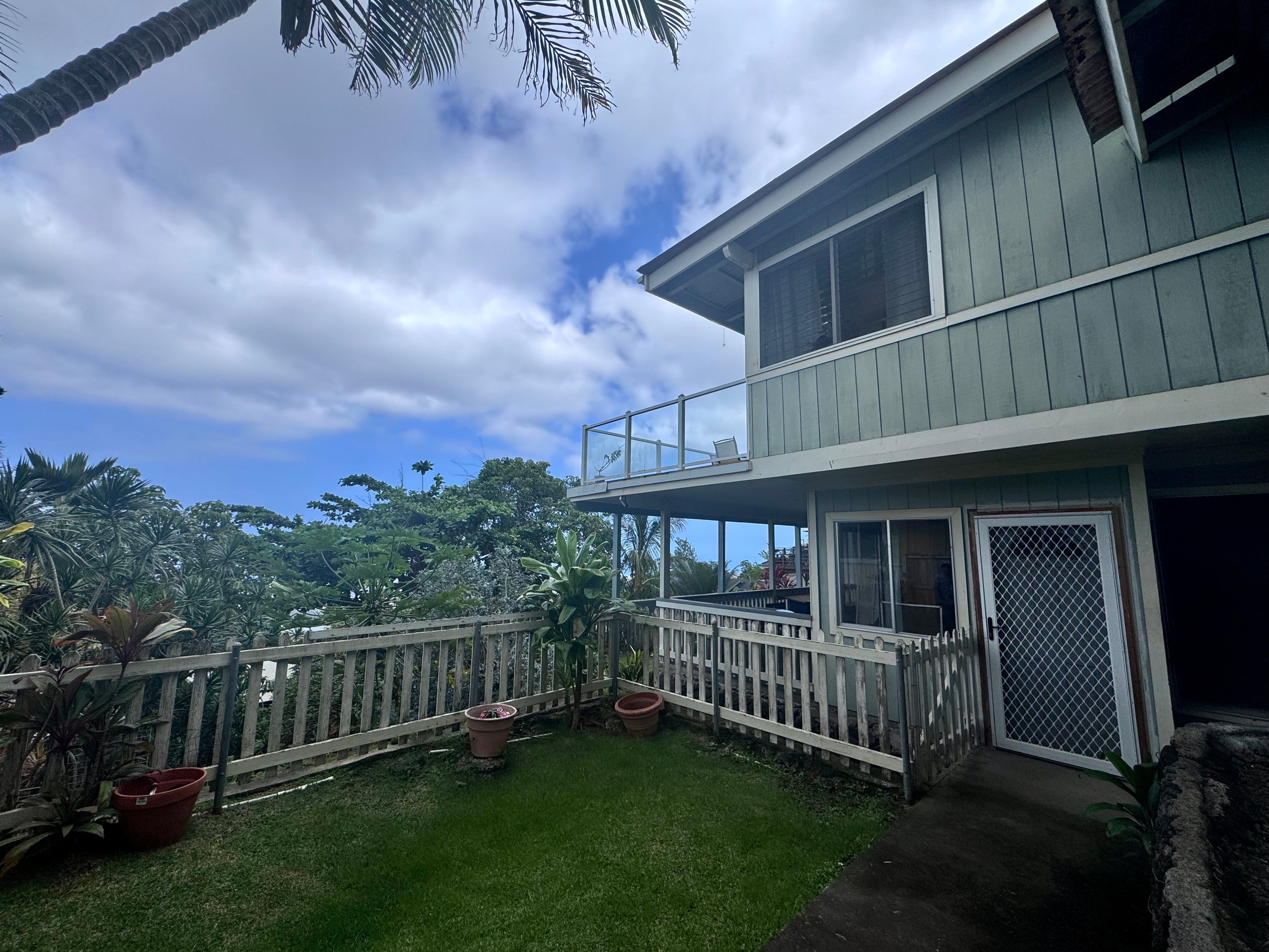 Two-story blue-gray house beside a fenced lawn and tropical plants, with a glass-railed upper balcony.