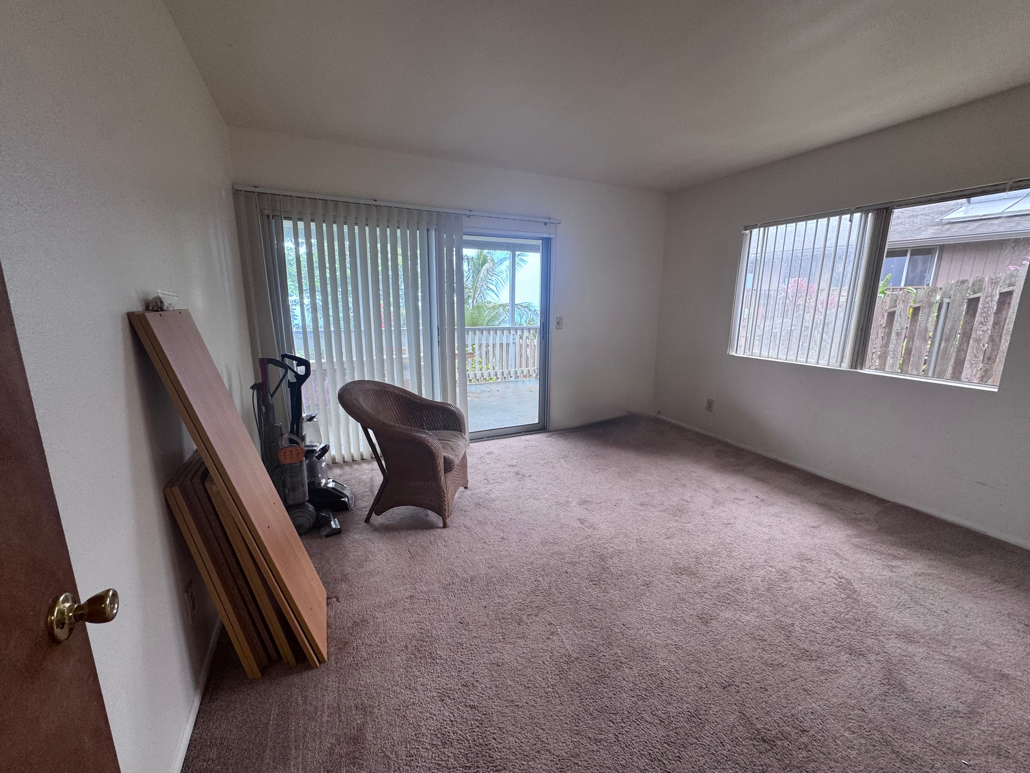 Carpeted room with a sliding glass door, vertical blinds, and a wicker chair beside stacked wooden panels.