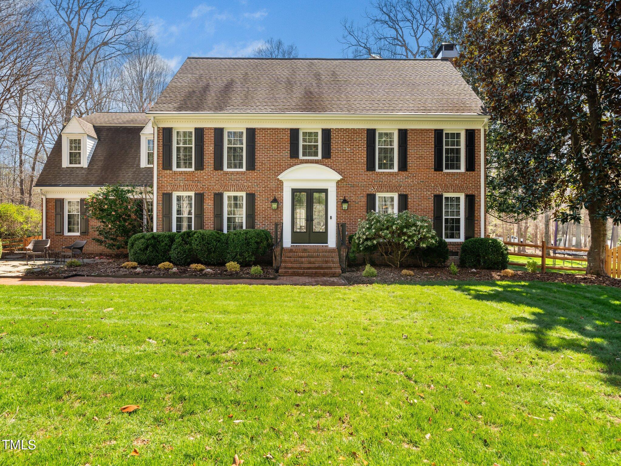 Two-story brick house with black shutters, landscaped shrubs, and a broad front lawn.