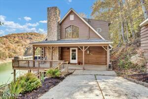 Wood-sided house with a stone chimney, covered porch, and raised deck beside a wooded hillside.