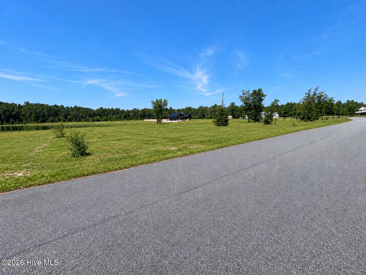 Grassy property beside a paved road, with a pond and homes in the distance.