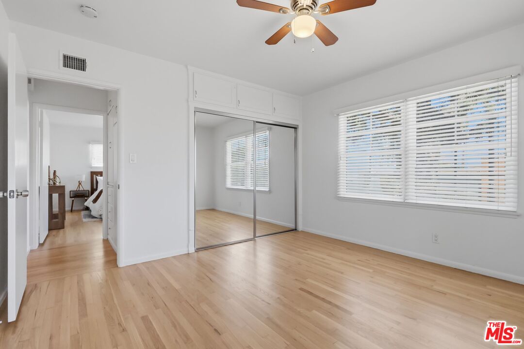 Bright empty bedroom with mirrored closet doors, hardwood floors, a ceiling fan, and wide windows with blinds.