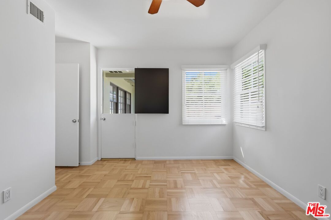 Empty bedroom with parquet flooring, two blind-covered windows, and a wall-mounted television.