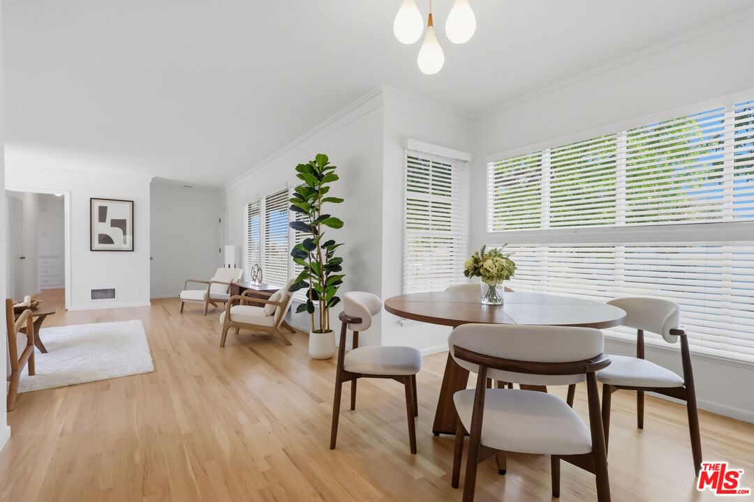 Staged dining area with a round wooden table, upholstered chairs, and large windows with white blinds.