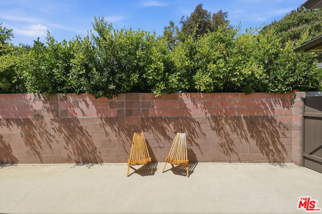 Concrete patio with two wooden folding chairs beside a block wall and tall hedges.