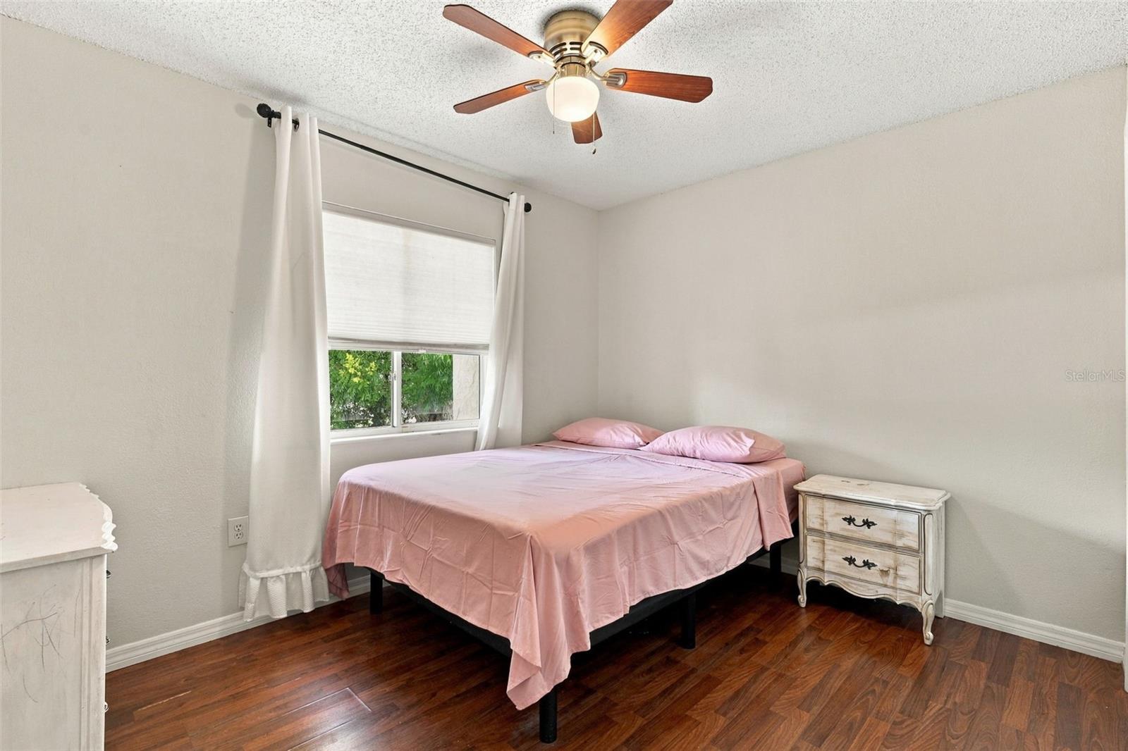 Bedroom with pink bedding, dark wood floors, a ceiling fan, and a curtained window.