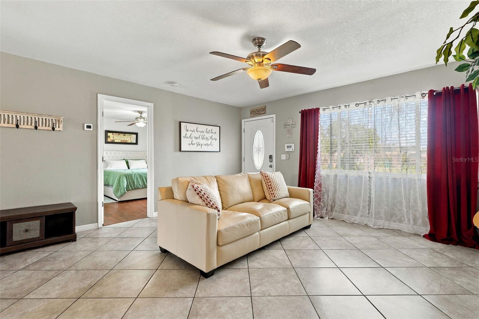 Living room with a cream leather sofa, tiled floor, ceiling fan, and burgundy curtains framing a large window.