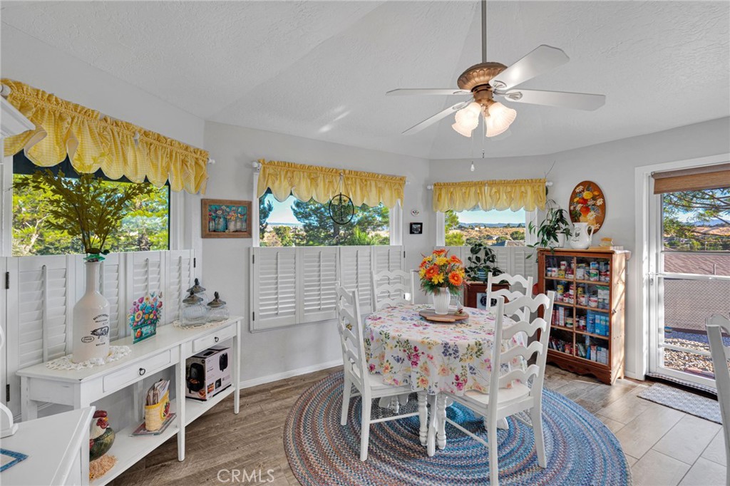 Dining room with a floral table, white chairs, plantation shutters, and windows overlooking trees and distant hills.