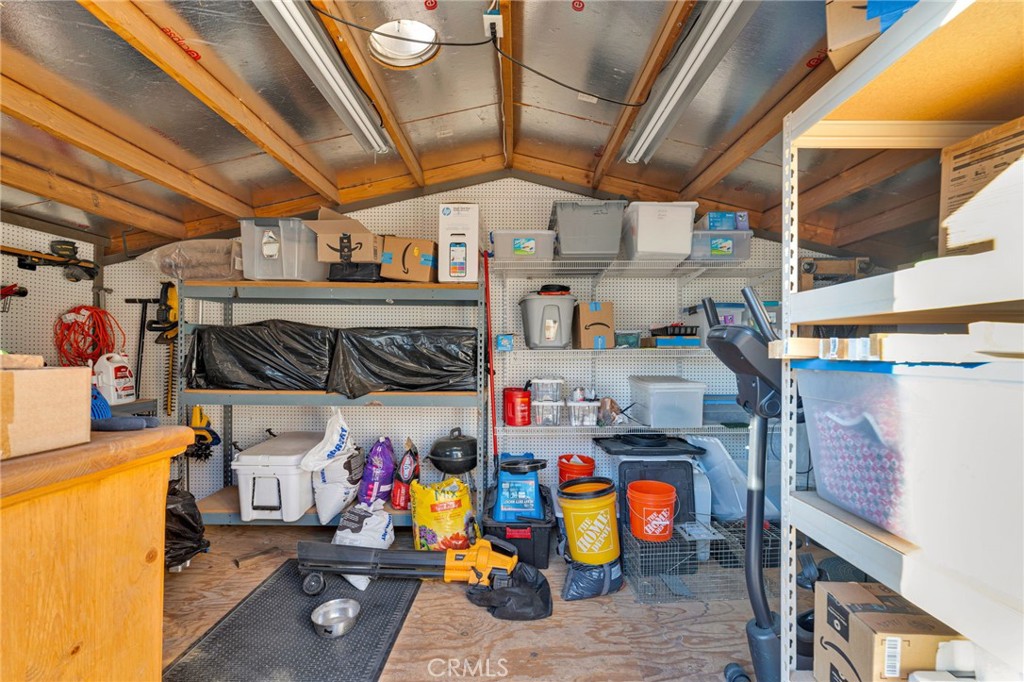 Storage shed filled with shelving, boxes, buckets, and household supplies beneath exposed wooden rafters.