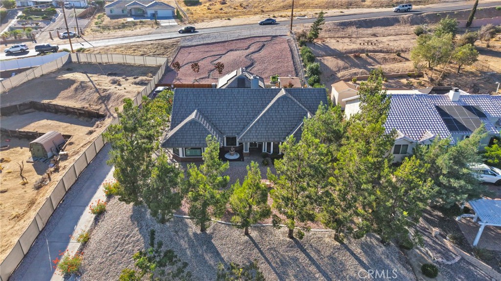 Aerial view of a dark-roofed home with a gravel yard, mature trees, and fenced lot.