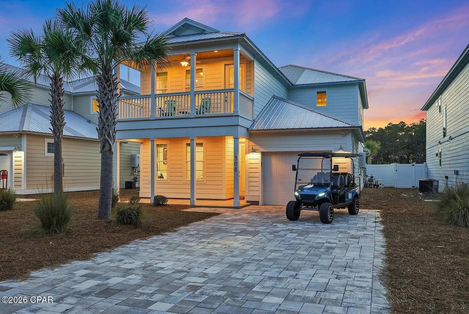 Two-story coastal-style home with a covered balcony, palm trees, and a golf cart in the driveway at dusk.
