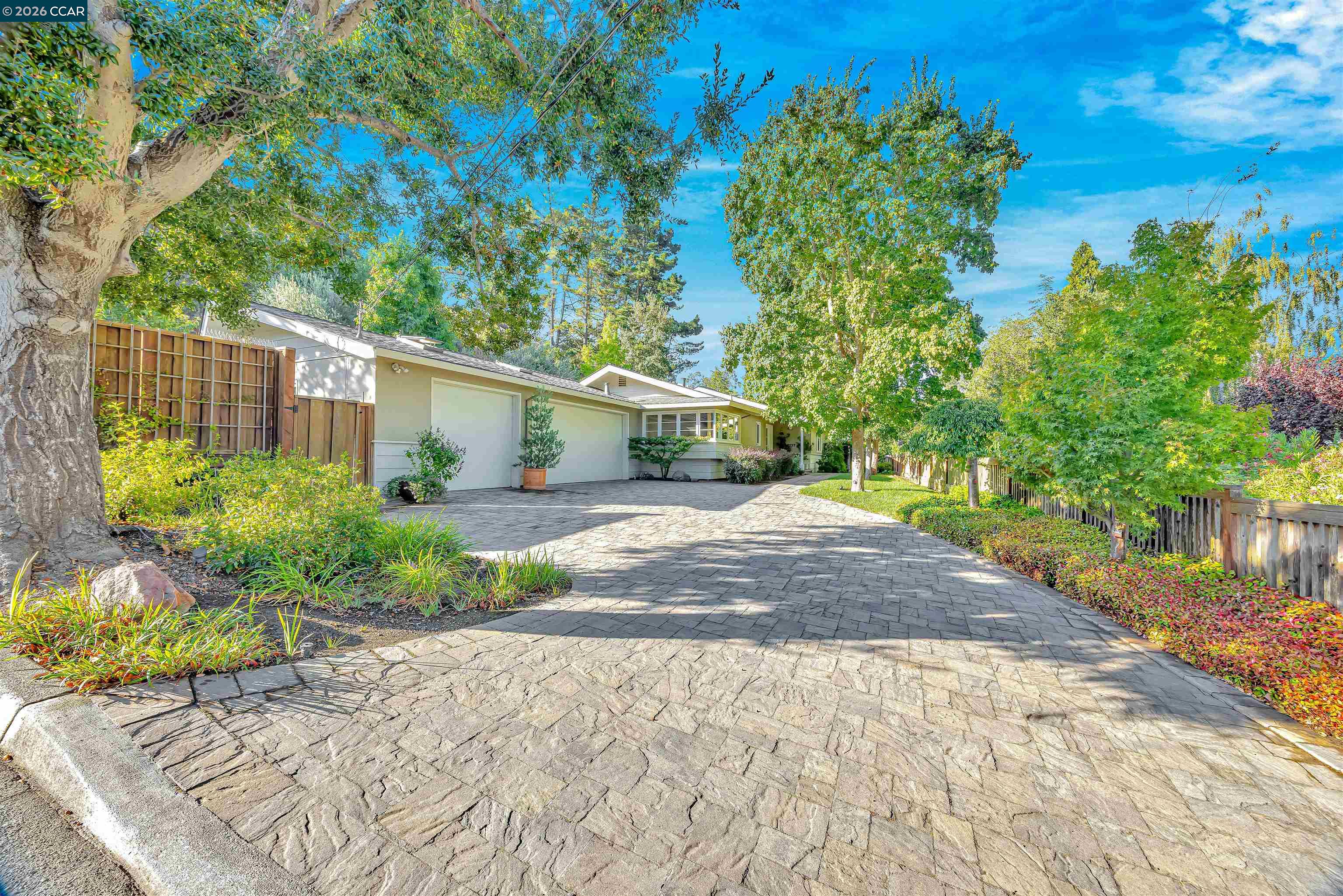Single-story home behind a broad stone-paver driveway, framed by mature trees and landscaped gardens.
