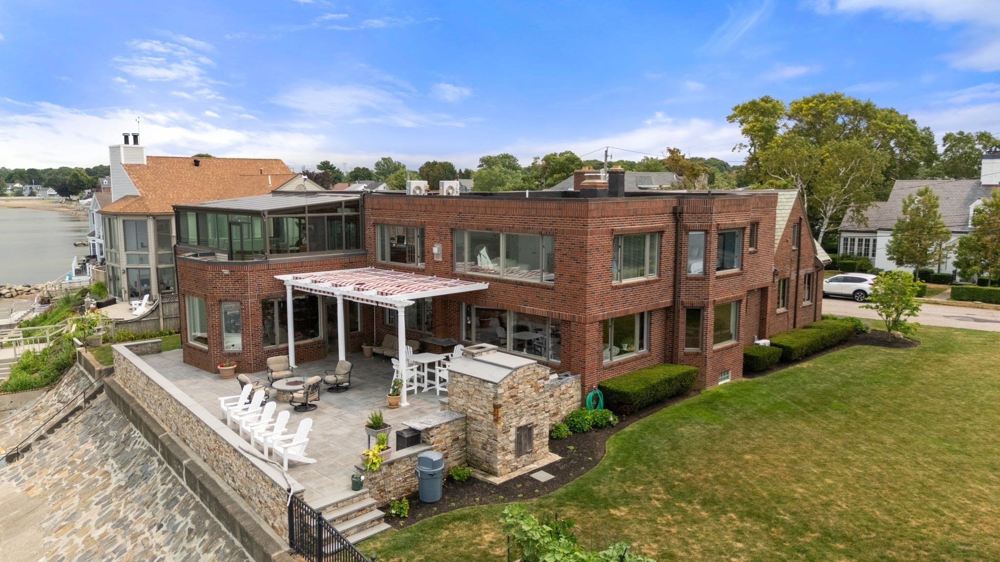 Brick waterfront home with a broad stone patio, white pergola, and seawall beside the water.