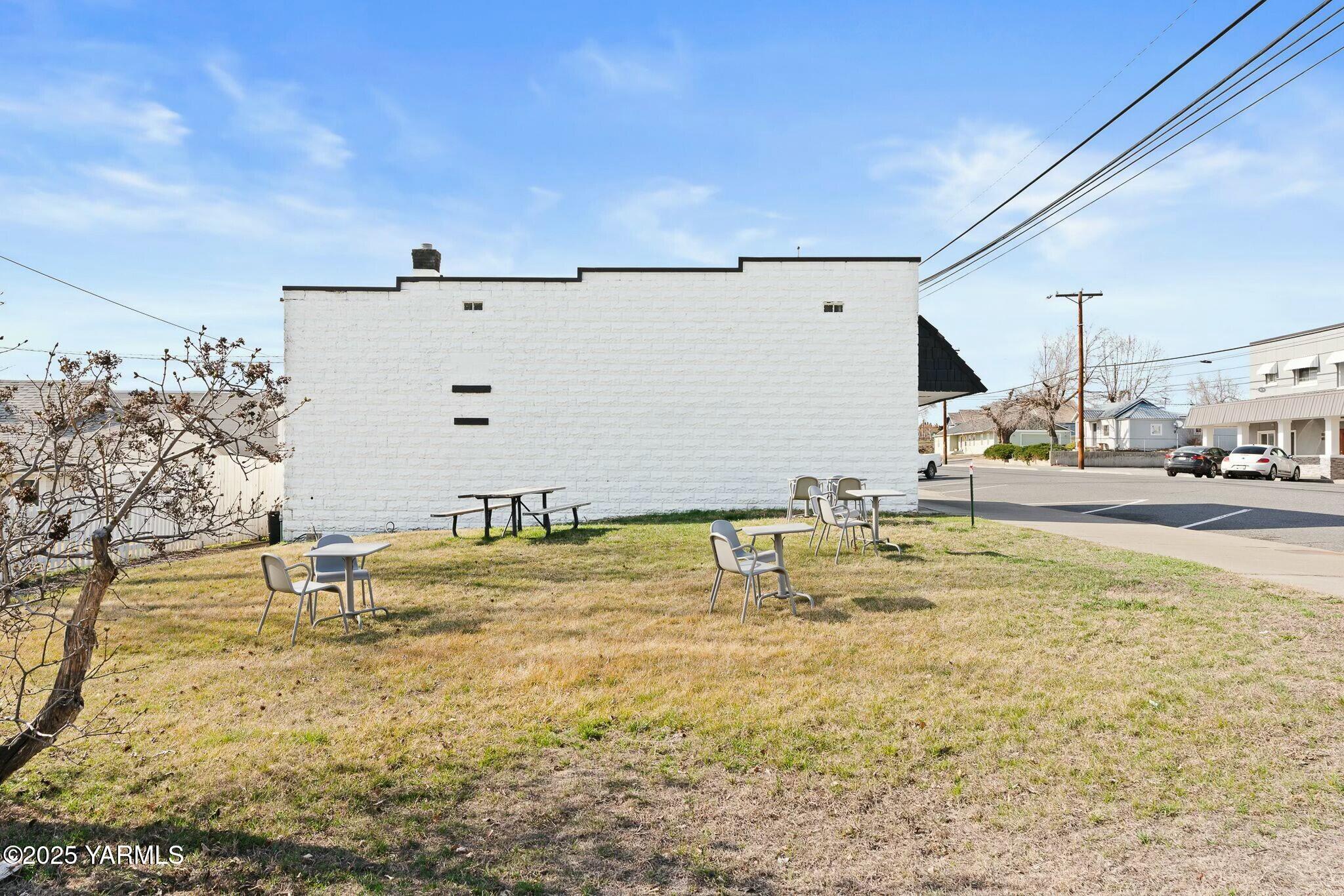 White masonry building beside a grassy outdoor seating area with picnic tables and chairs.