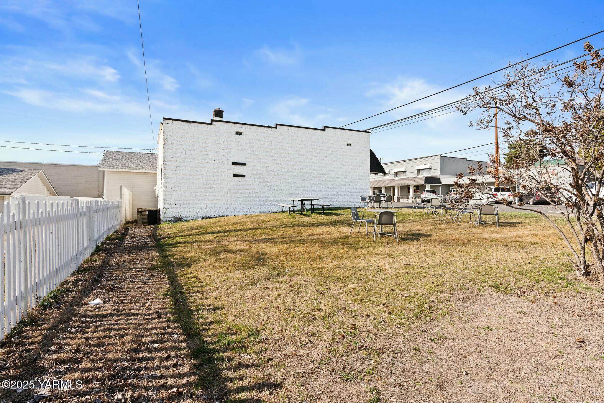Grassy yard beside a white masonry building, with a picket fence and scattered outdoor seating.