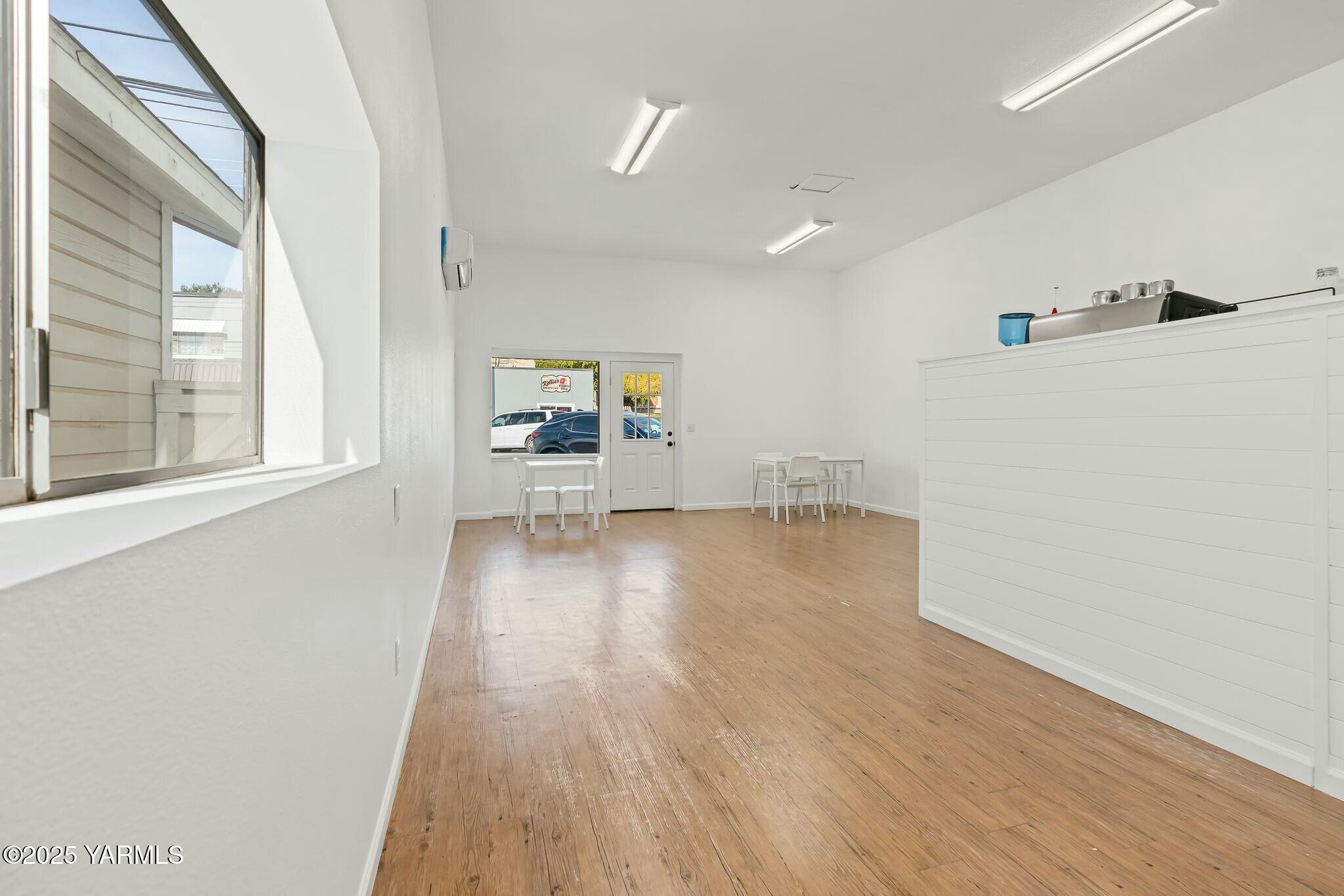 Bright commercial interior with wood flooring, a white service counter, espresso machine, and small tables.