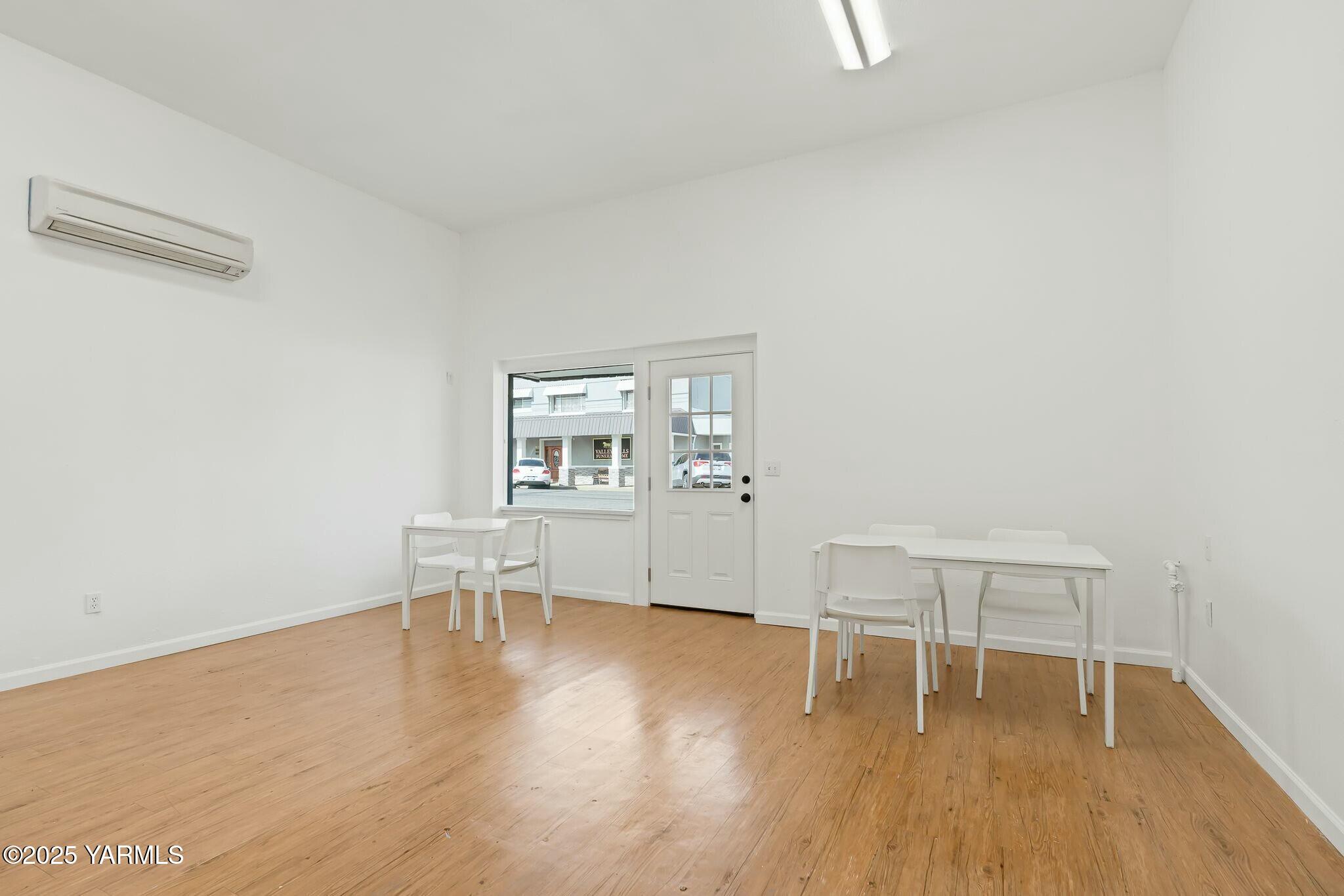 Bright room with wood flooring, white tables and chairs, and a glass-paneled door beside a window.