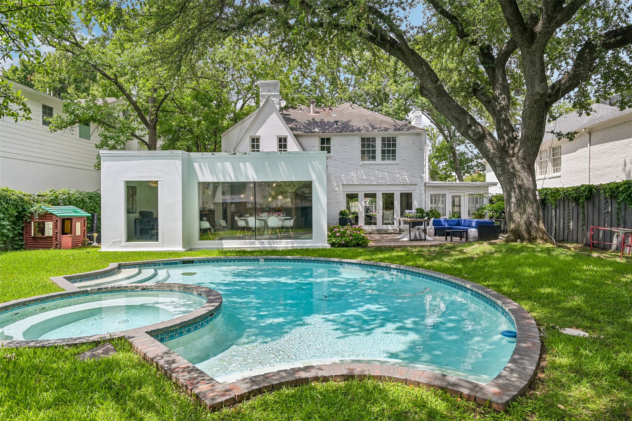 White house with glass-walled extension behind a curved swimming pool and patio beneath mature trees.