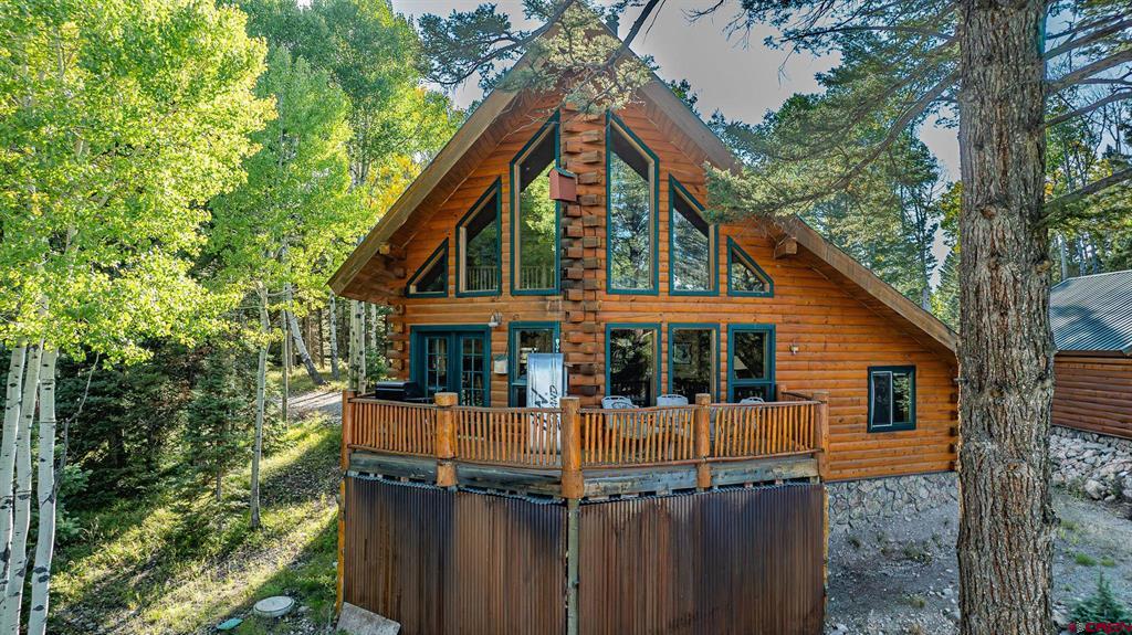 Log cabin with a steep A-frame roof and wooden deck among mature trees.