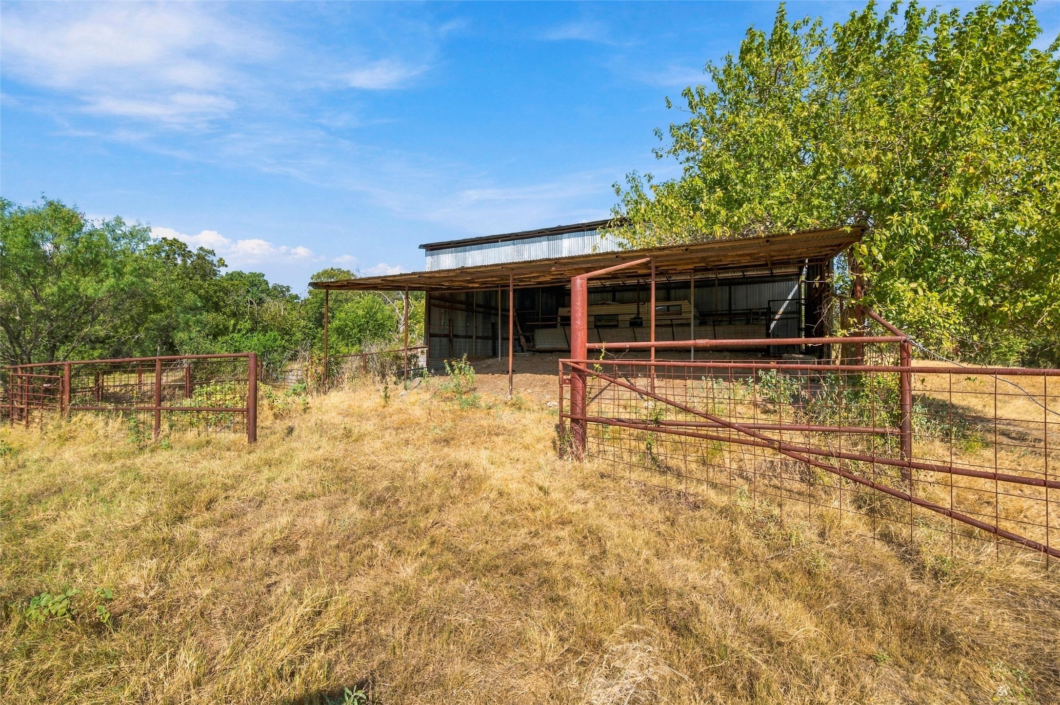 Rural property with a metal livestock shelter, rust-colored fencing, and mature trees.