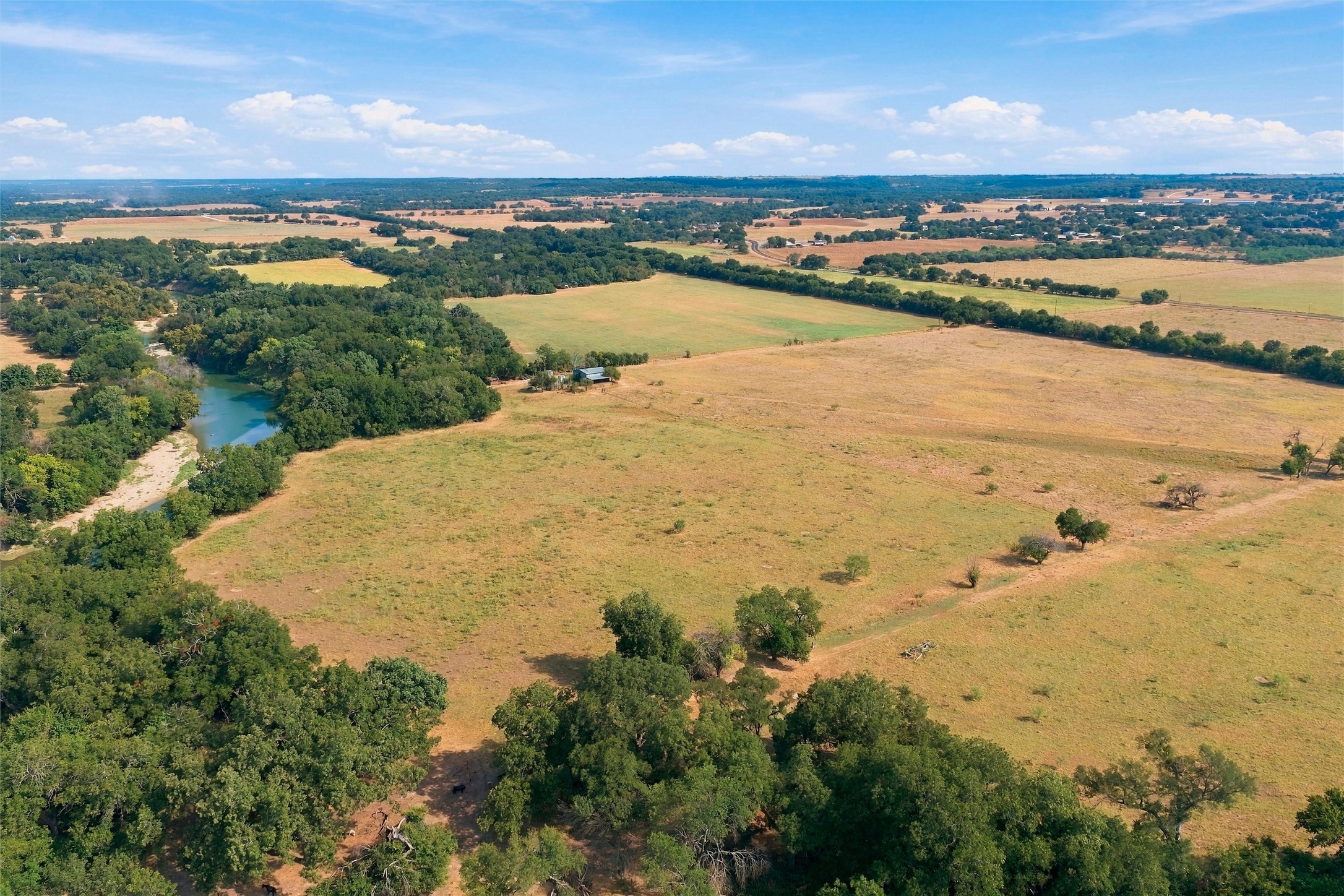 Aerial view of open ranchland with a wooded creek, scattered trees, and small farm buildings.