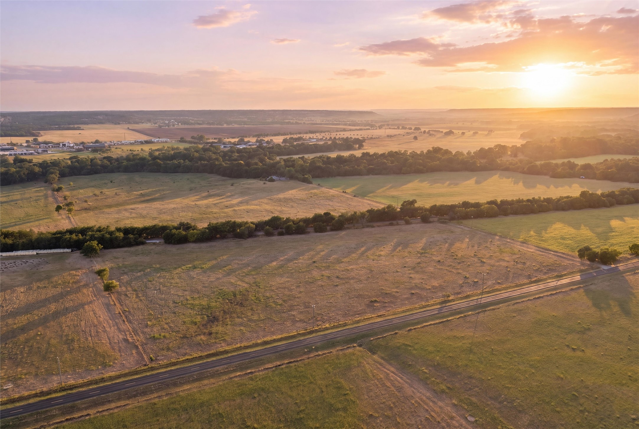 Aerial view of open acreage and fields beside a road at sunset, with tree lines across the horizon.