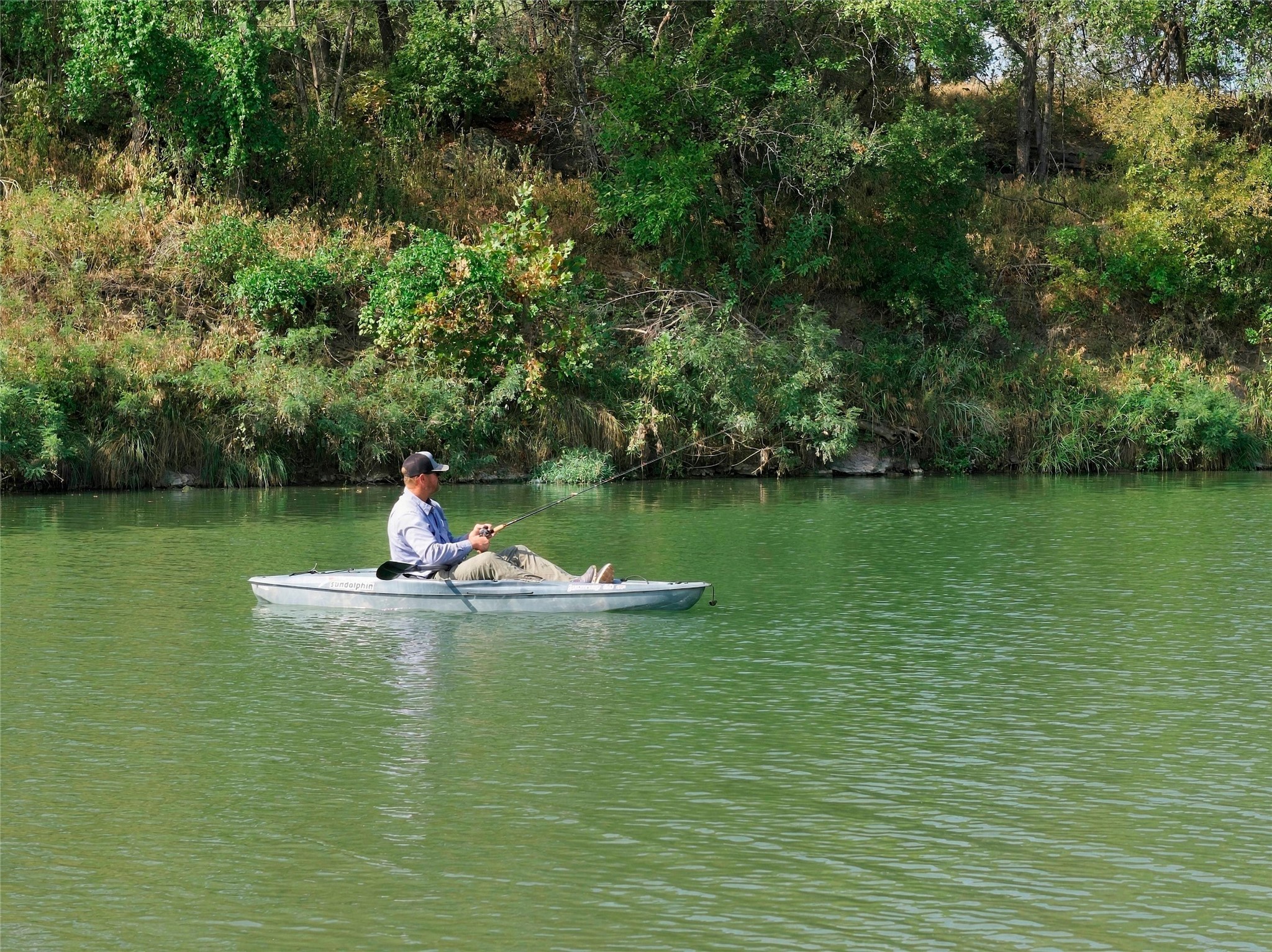 A man fishes from a small kayak on green water beside a wooded bank.