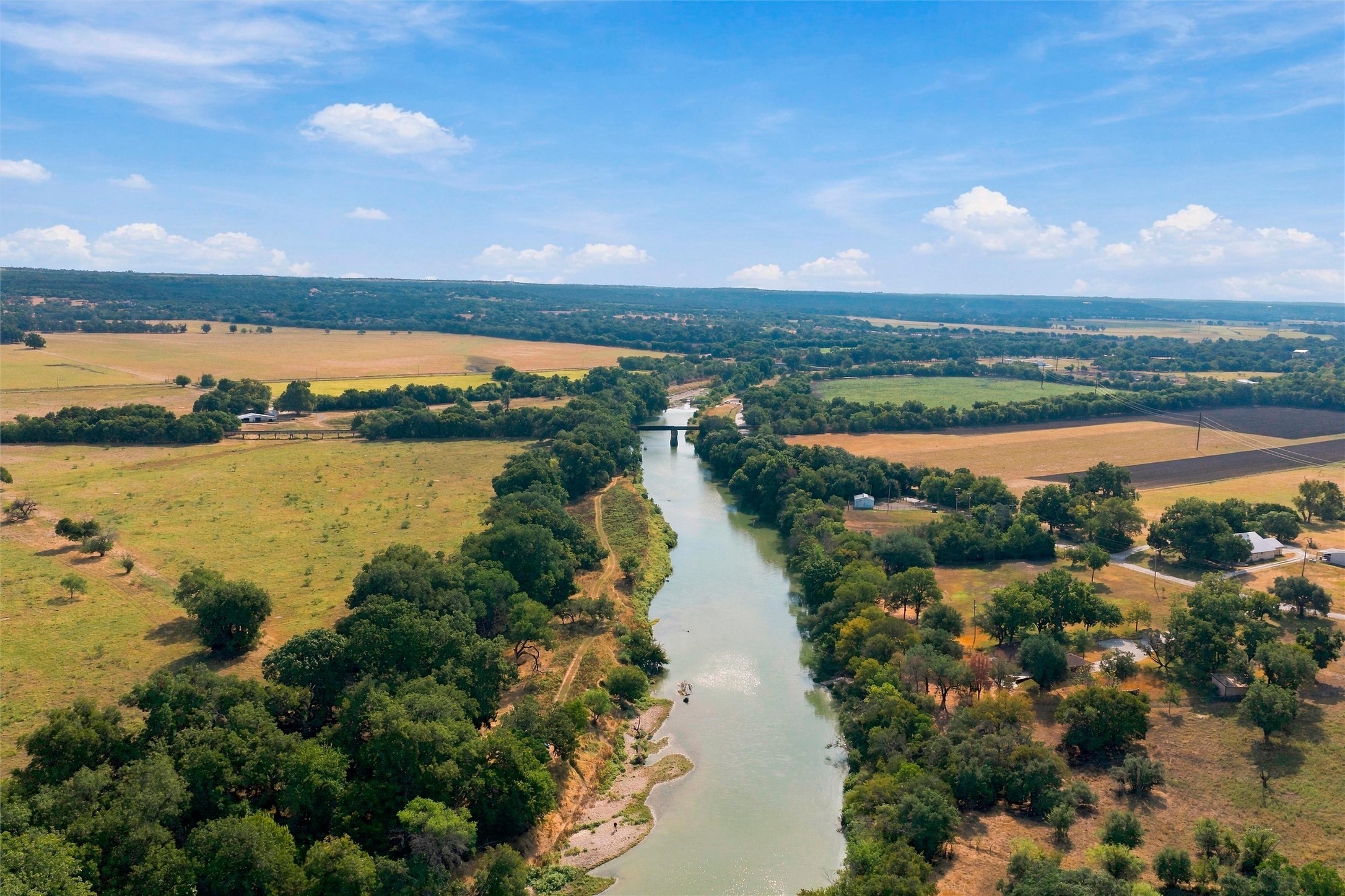 Aerial view of a tree-lined river winding through open fields, with a bridge in the distance.