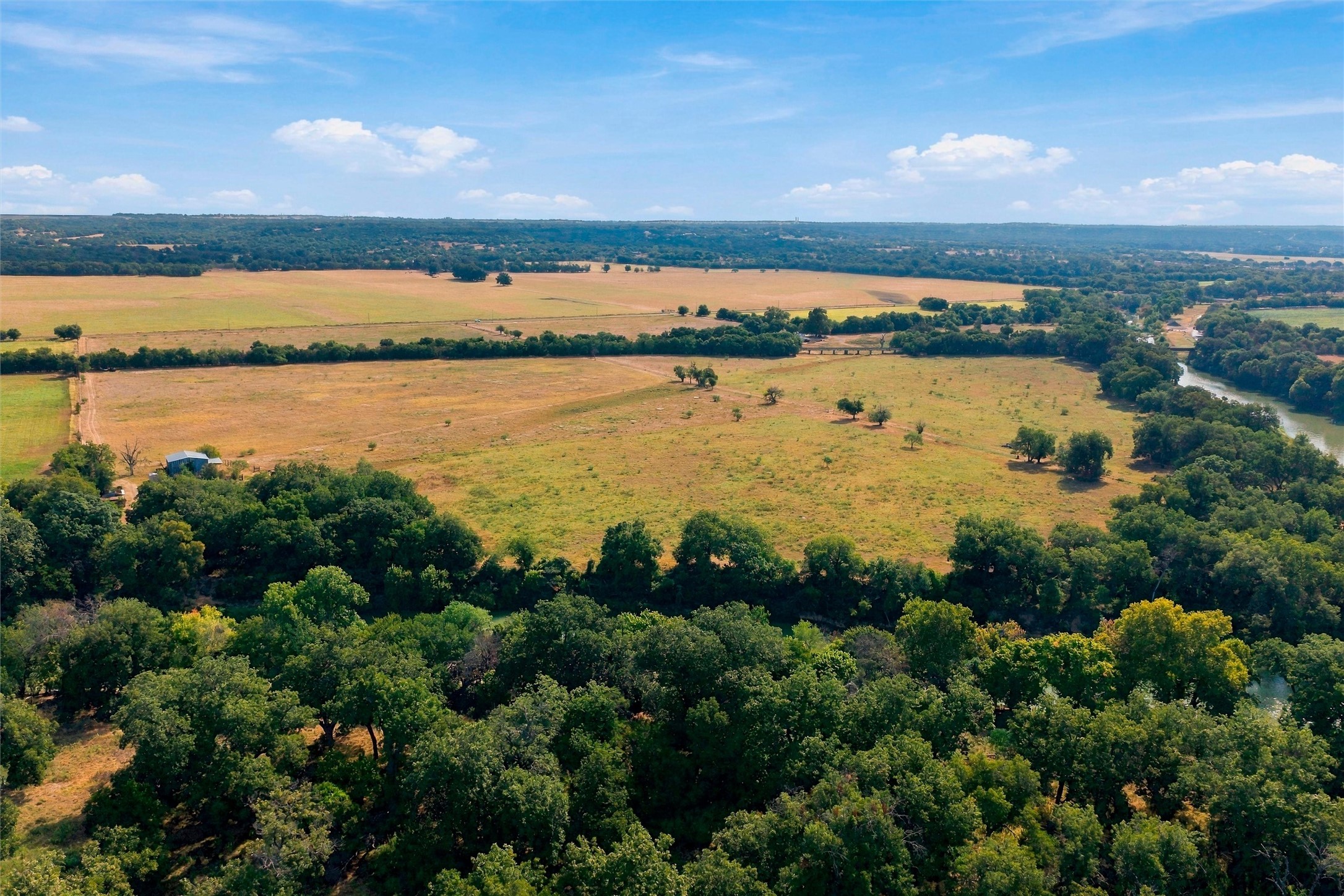 Aerial view of open acreage with broad fields, wooded edges, a small outbuilding, and a river.