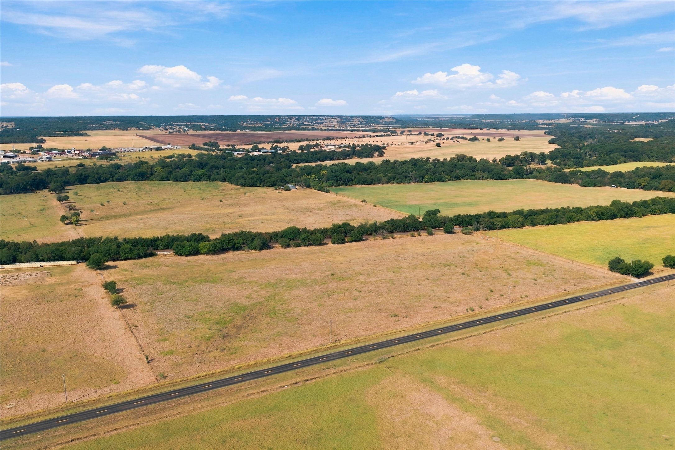 Aerial view of open rural fields bordered by trees, with a paved road across the foreground.