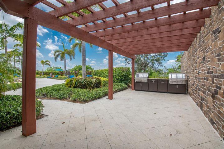 Covered patio with a wood pergola, stone wall, and built-in outdoor kitchen beside tropical landscaping.