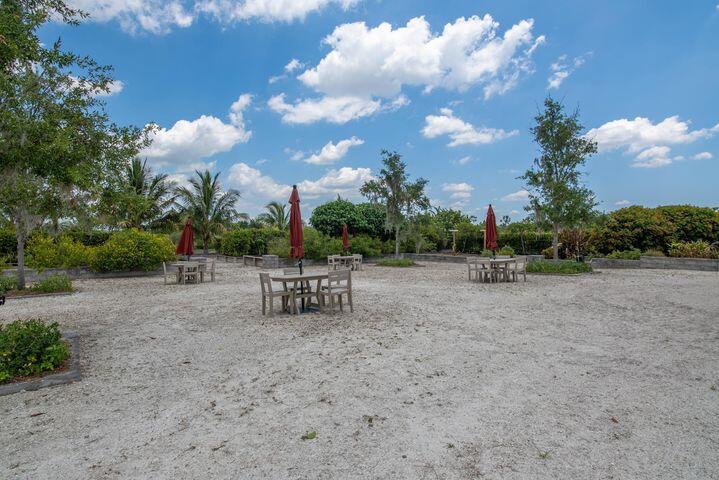 Gravel outdoor seating area with wooden tables and closed red umbrellas, bordered by trees.