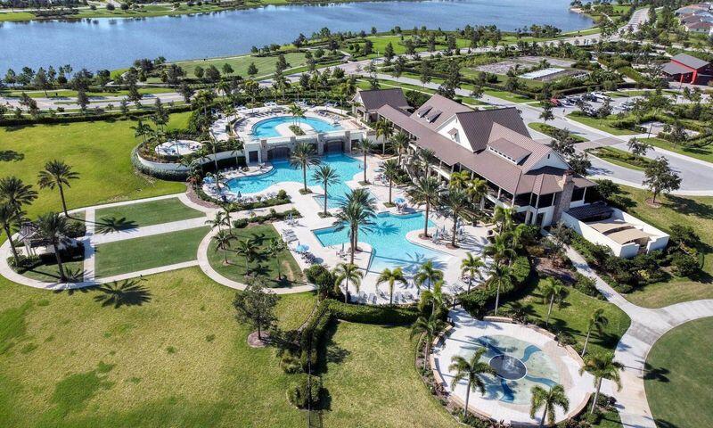 Aerial view of a clubhouse and swimming pools beside a lake, surrounded by palm trees and landscaped paths.