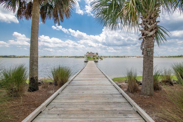 Wooden dock extends across a lake toward a small covered structure, framed by palm trees.