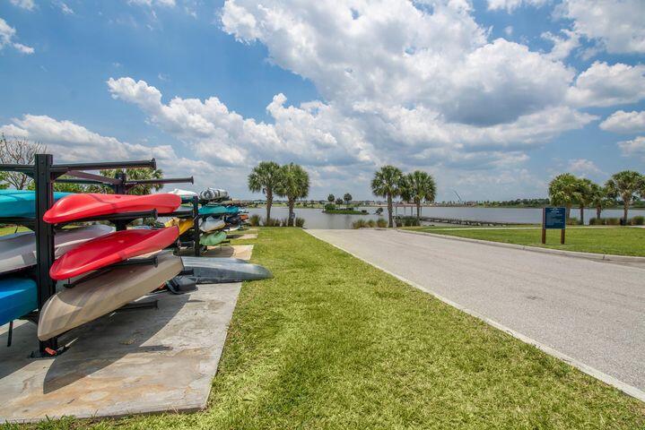 Kayaks on racks beside a grassy lakeside road and palm-lined water.