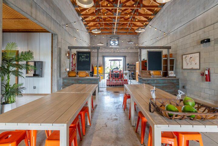 Rustic dining area with long wooden tables, orange stools, and an exposed timber-truss ceiling.