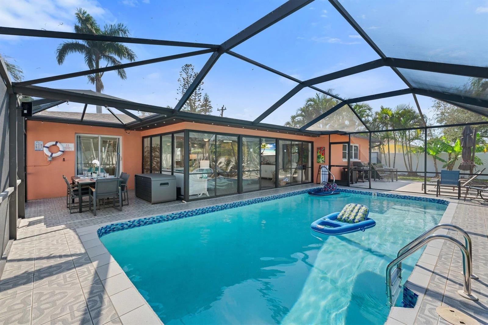 Screened-in swimming pool beside a peach-colored home, with a patio dining area and floating loungers.