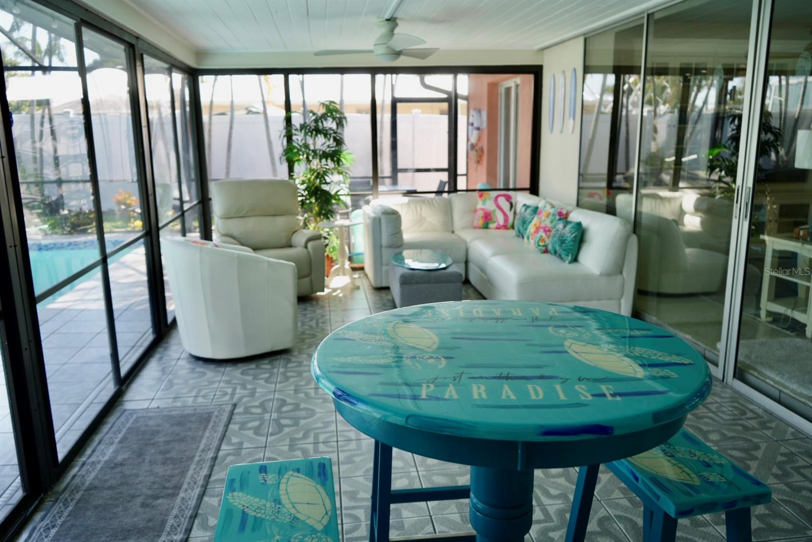 Glass-enclosed sunroom with white seating and a turquoise table overlooking the pool.