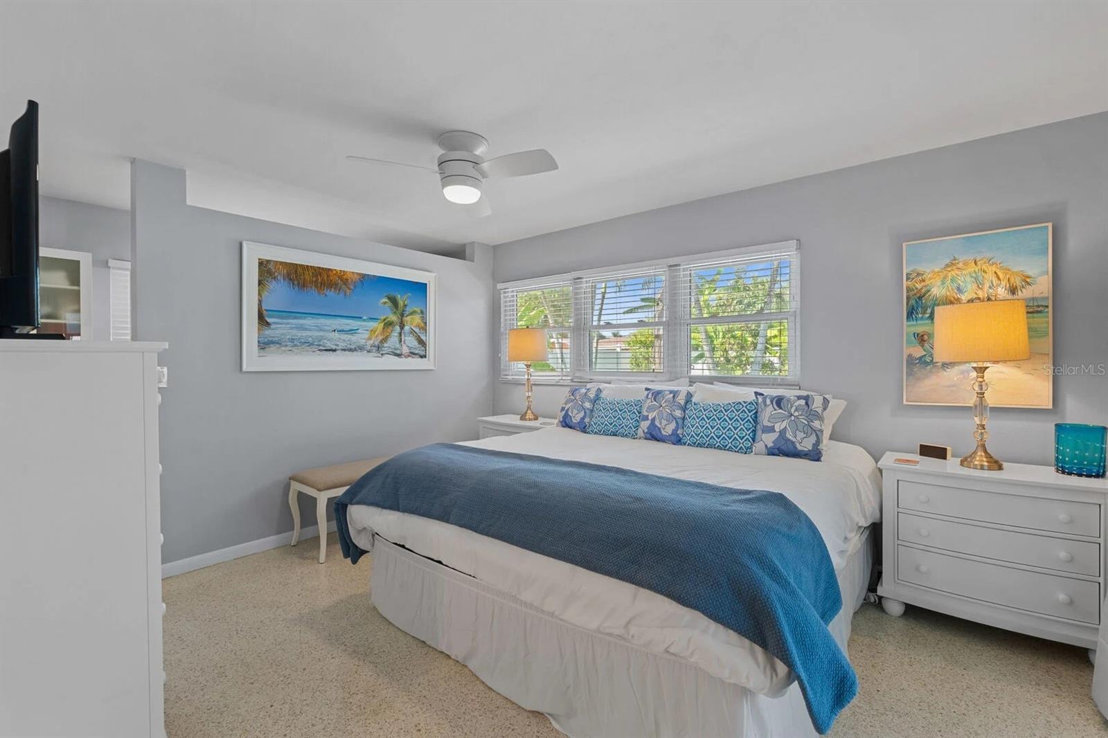Furnished bedroom with a large bed, blue accents, ceiling fan, and wide windows.