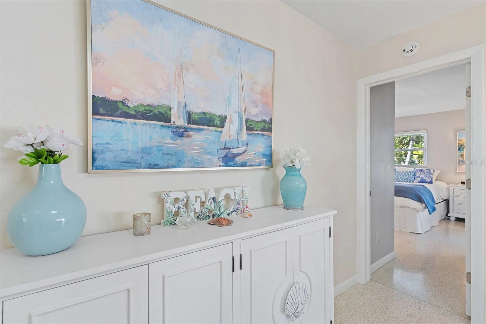 Hallway with a white console cabinet, turquoise vases, and sailboat painting beside an open bedroom doorway.
